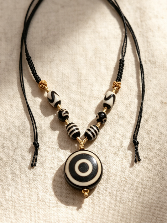 A black cord necklace with a central round pendant and striped beads, laid flat on a textured beige fabric background.