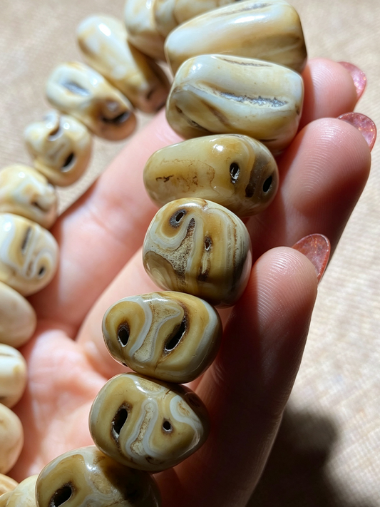 A close-up of a hand holding a strand of carved, polished beads with swirling natural patterns in cream, tan, and brown.