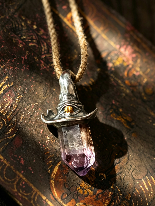 A witch hat pendant necklace made of silver-toned metal with a purple crystal point and a small gold eye detail, hanging from a tan cord against an ornate dark book background.