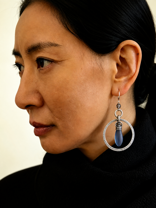 A close-up side profile of a woman wearing a large silver-toned dangle earring with a dark blue teardrop-shaped stone pendant.