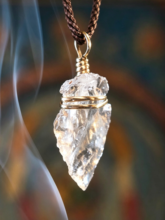 A clear crystal point pendant necklace with gold wire wrapping, suspended from a brown braided cord, shown against a blurred warm-toned background with a smoky white streak on the left.