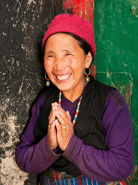 A smiling woman wearing a bright pink knit beanie, a purple long-sleeve top, and a black vest, standing against a weathered dark wall and a green door.She was wearing Tibetan earrings and a necklace.
