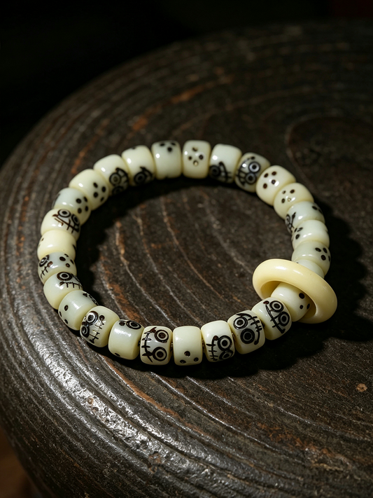 A beaded stretch bracelet made of cream-colored bone or ivory beads, featuring black skull and dot patterns, with a large solid cream disc bead, displayed on a dark wooden surface.