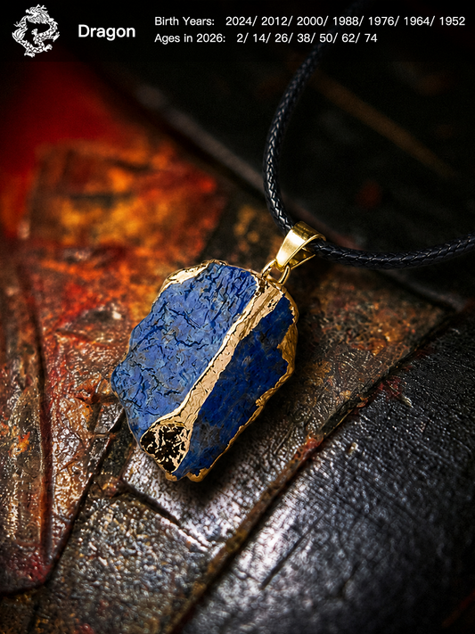 A raw blue lapis lazuli crystal pendant necklace with gold-colored edges and a black cord, displayed on a dark textured surface with a warm, rustic background. White text in the top left corner reads 'Dragon' with birth years and ages in 2026.