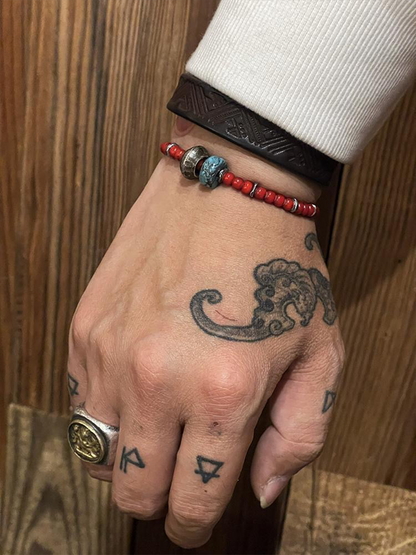 A close-up of a hand wearing a red beaded bracelet with a blue-green bead accent and a dark brown leather wristband, against a wooden background.