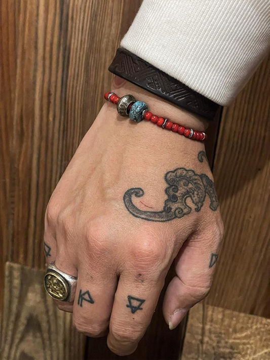 A close-up of a hand wearing a red beaded bracelet with a blue-green bead accent and a dark brown leather wristband, against a wooden background.