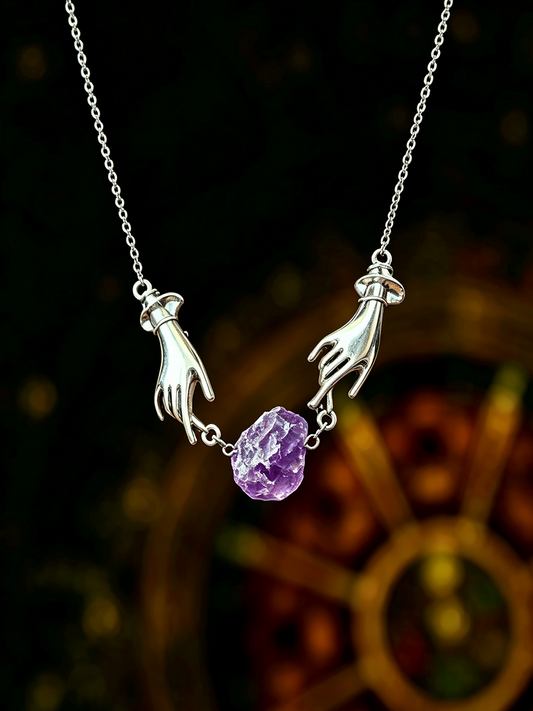 A silver pendant necklace featuring two hands reaching toward a faceted purple crystal, suspended from a delicate silver chain against a dark, blurred background.