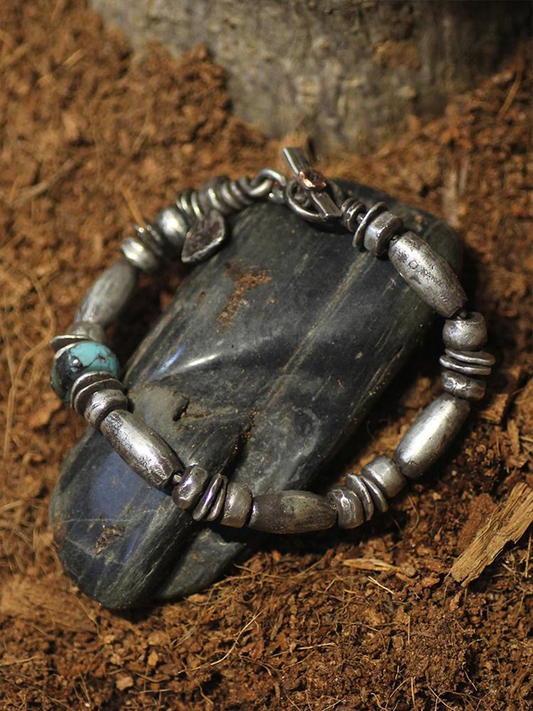 A handmade silver beaded bracelet with a small turquoise accent, displayed on a dark polished stone over a brown soil background.
