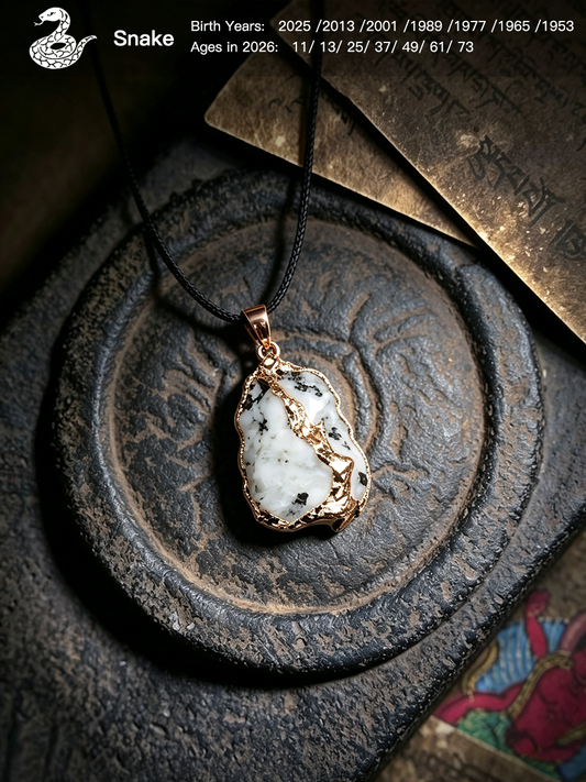 A snake pendant necklace with a polished white stone featuring black and gray marbling, set in a textured rose gold or copper-toned bezel. The pendant hangs from a black cord on a dark, rustic circular stone plate with a gold-toned card in the background.