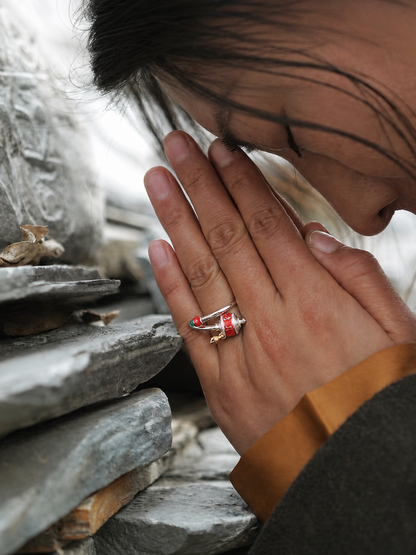 A close-up lifestyle shot of a person with hands in prayer pose, wearing a silver-toned ring with red and gold Tibetan prayer flag details on the ring finger, set against a background of stacked gray stone tablets.