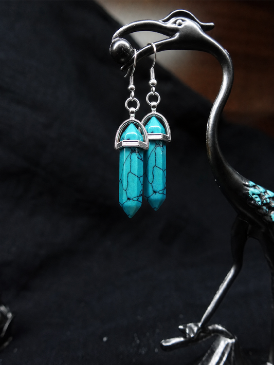 A pair of turquoise crystal point dangle earrings with silver metal caps and hooks, hanging from a black metal bird-shaped jewelry stand against a dark fabric background.
