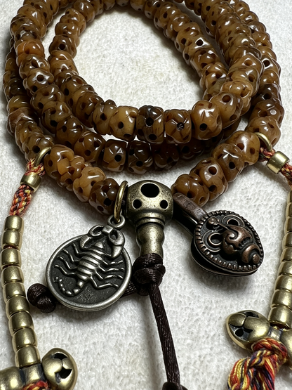Brown beaded necklace with skull-shaped beads, antique bronze and silver-toned metal charms, and braided cord tassels on a white textured background.