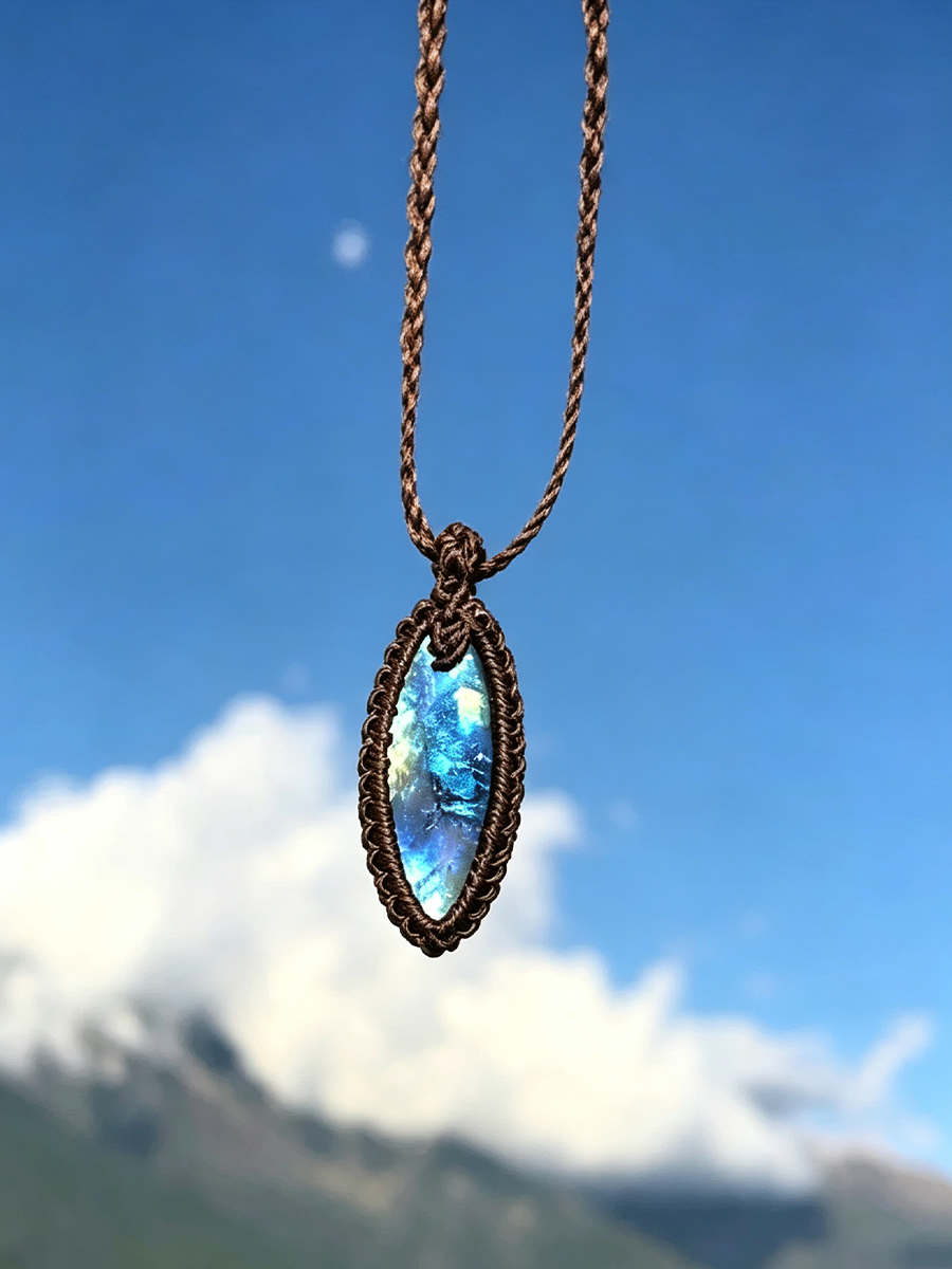 A handmade oval pendant necklace with a blue iridescent abalone shell in a dark brown woven basket setting, hanging against a blurred background of blue sky and white clouds.