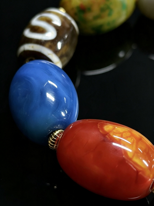 Close-up of glossy oval lampwork beads on a dark surface, including a prominent blue bead and a red-orange bead with yellow marbling, with other beads blurred in the background.