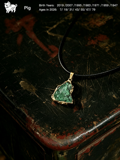 A raw green stone pendant necklace with a gold-toned bezel and black cord, displayed on a distressed dark wooden surface. The image includes a Pig logo and birth/age details in the top left corner.