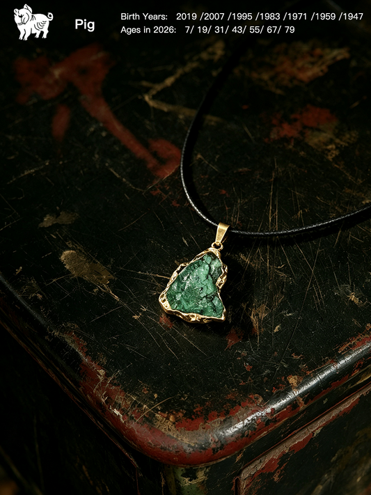 A raw green stone pendant necklace with a gold-toned bezel and black cord, displayed on a distressed dark wooden surface. The image includes a Pig logo and birth/age details in the top left corner.