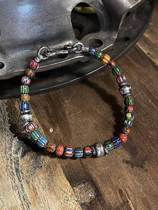 A handmade beaded bracelet with colorful striped glass beads and silver-toned metal spacers, displayed on a rustic wooden surface next to a dark metal object.