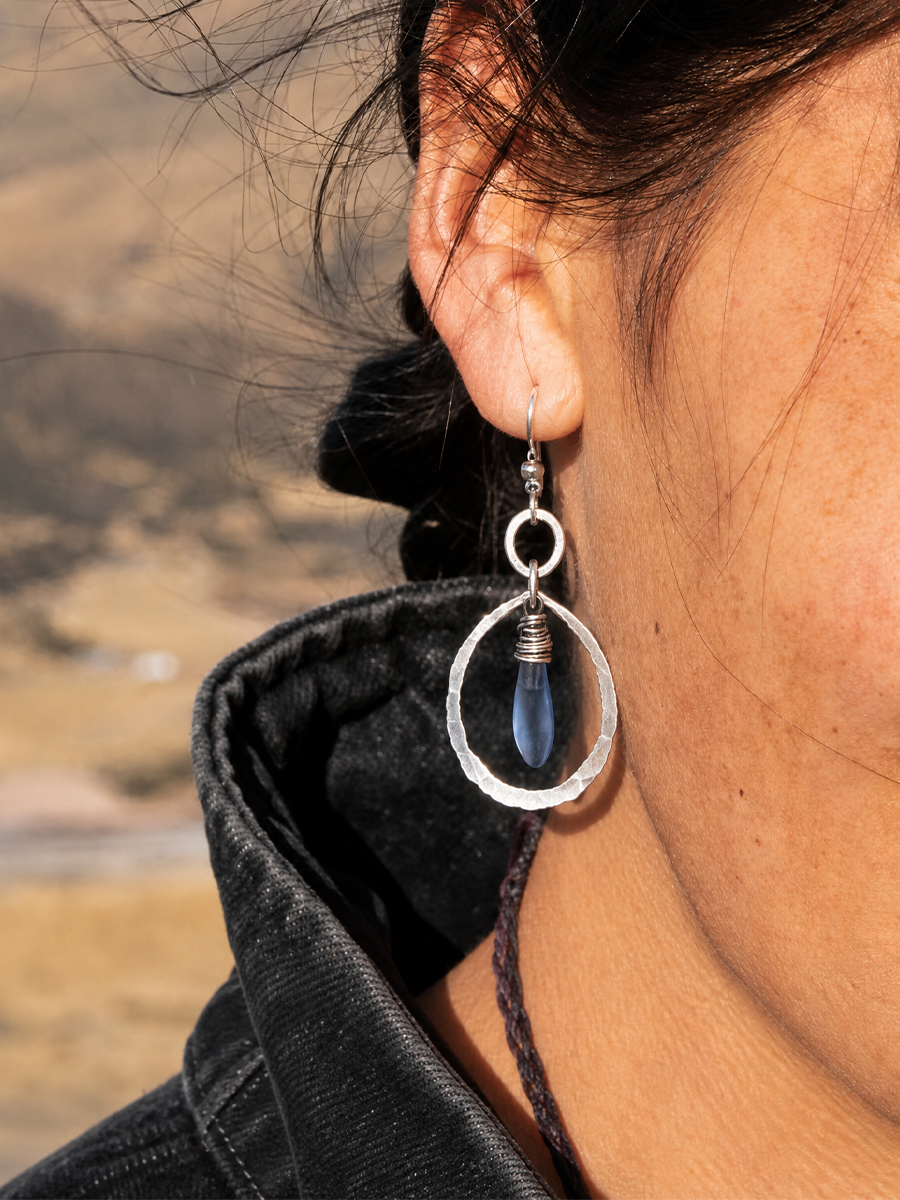 A close-up of a person's ear wearing a silver dangle earring with a textured silver oval frame and a dark blue teardrop bead, set against a blurred outdoor background.