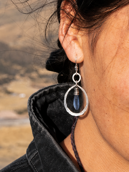 A close-up of a person's ear wearing a silver dangle earring with a textured silver oval frame and a dark blue teardrop bead, set against a blurred outdoor background.