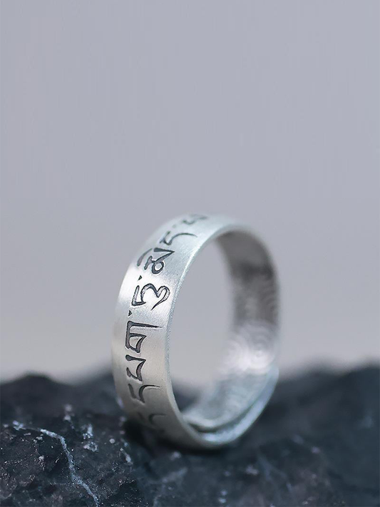 A silver-toned metal ring engraved with Tibetan script, resting on a dark textured stone surface against a soft gray background.