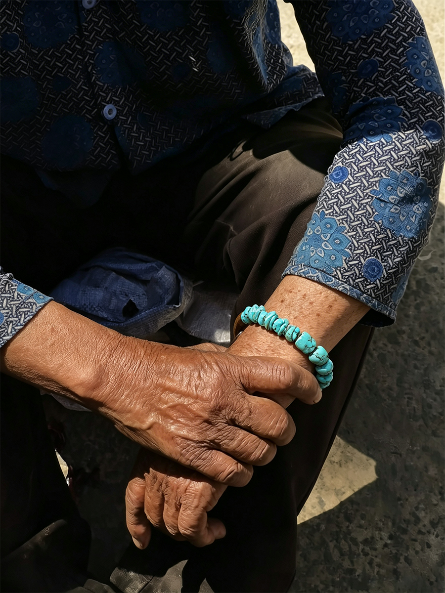 A close-up of an elderly person's weathered hands resting on their lap, wearing a turquoise beaded bracelet on one wrist. The person is wearing a blue and black patterned long-sleeve shirt and dark trousers, seated outdoors on a concrete surface.
