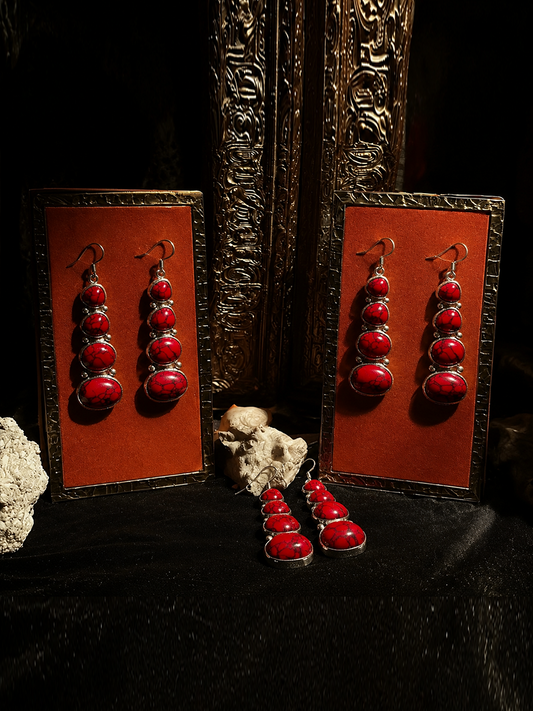 A pair of long drop earrings with oval red stones in silver settings, displayed on two orange card holders with additional matching earrings placed in front, against a dark ornate background.