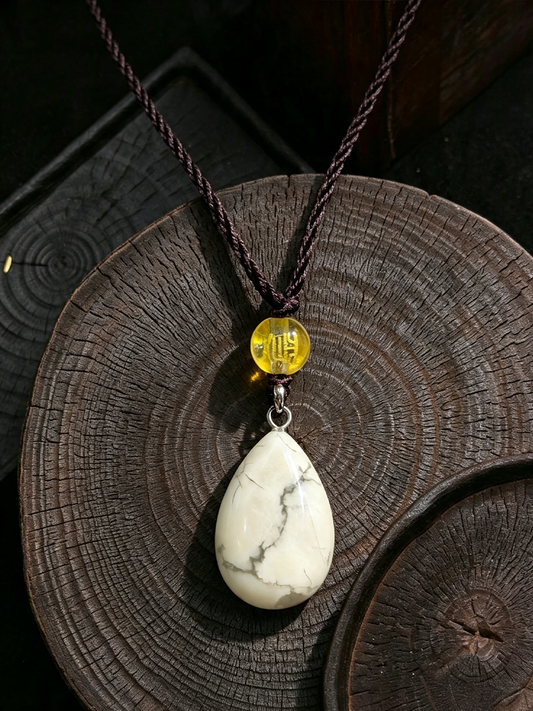 A teardrop-shaped white stone pendant necklace with natural gray veining, hanging from a dark brown braided cord and a small round yellow accent bead, displayed on a dark wood slice background.