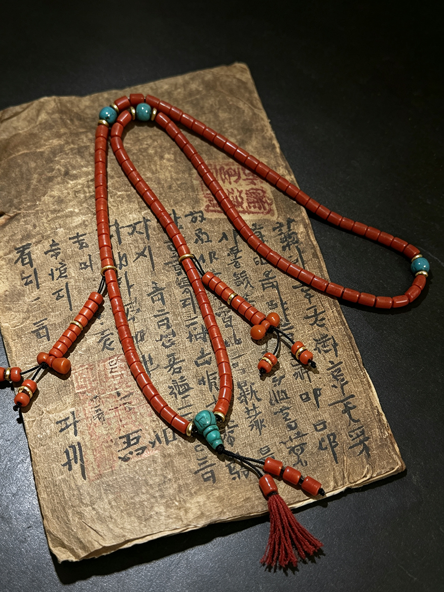 A red beaded necklace with turquoise-colored beads and a red tassel, draped over an aged, handwritten paper document on a dark surface.