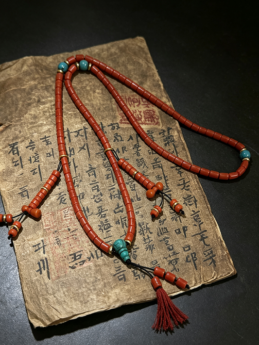 A red beaded necklace with turquoise-colored beads and a red tassel, draped over an aged, handwritten paper document on a dark surface.
