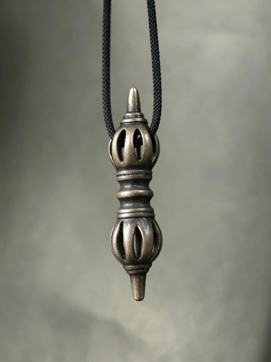 An antique bronze-toned metal pendant with intricate cutout details hangs from a black braided cord against a soft gray background.