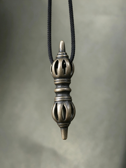 An antique bronze-toned metal pendant with intricate cutout details hangs from a black braided cord against a soft gray background.