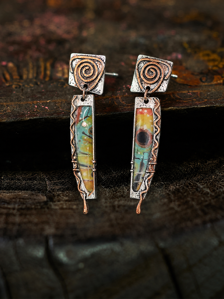 A pair of handmade dangle earrings with silver-toned metal frames, copper-toned spiral details, and colorful marbled resin inserts, displayed on a dark rustic wooden surface.