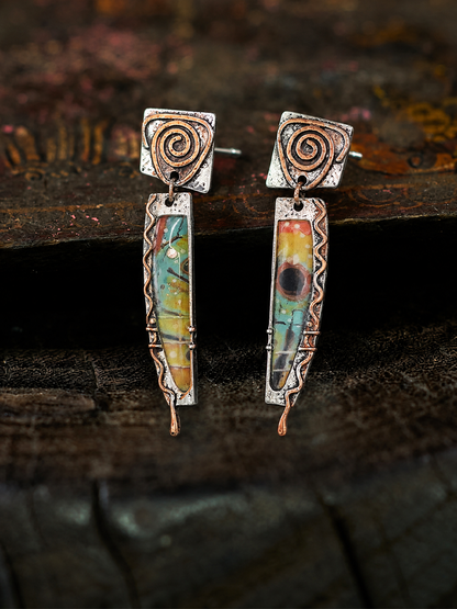 A pair of handmade dangle earrings with silver-toned metal frames, copper-toned spiral details, and colorful marbled resin inserts, displayed on a dark rustic wooden surface.