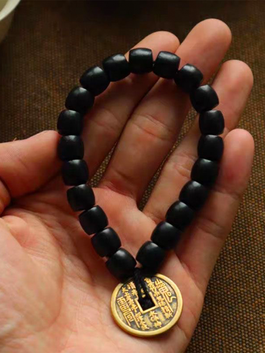 A black beaded bracelet with a gold-colored Chinese ancient coin pendant, held in a person's hand.
