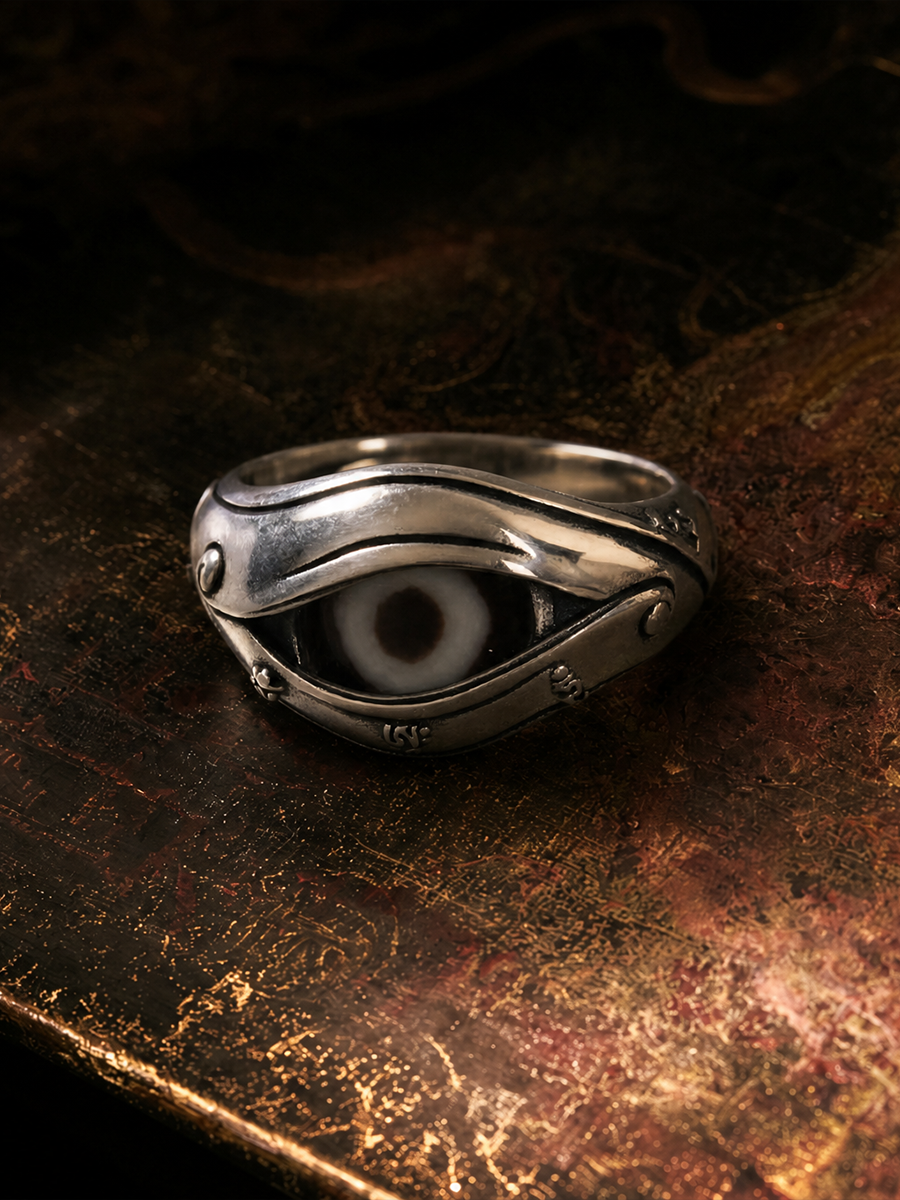 A silver-toned gothic ring featuring a stylized eye motif with a white and black iris, resting on a textured, dark metallic surface.