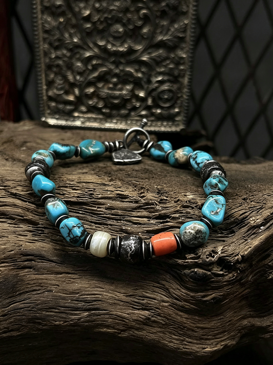 A beaded bracelet with turquoise blue, black, white, and orange beads, displayed on a piece of rustic wood with a decorative metal plaque in the background.