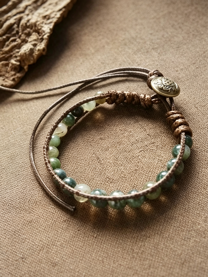 A bohemian-style multi-strand bracelet featuring green and white marbled beads on brown cord, with a small round metallic charm, displayed on a textured beige fabric background.