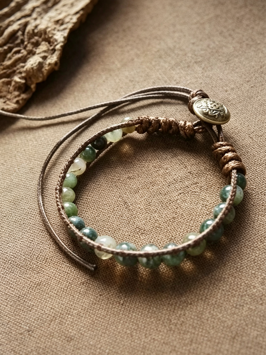 A bohemian-style multi-strand bracelet featuring green and white marbled beads on brown cord, with a small round metallic charm, displayed on a textured beige fabric background.