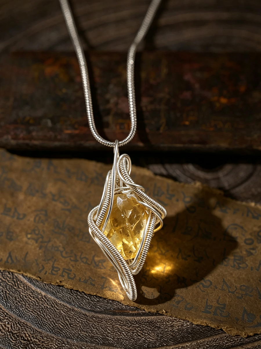 A silver wire-wrapped pendant necklace featuring a faceted yellow gemstone, displayed on a textured brown surface with a piece of aged, script-covered paper.