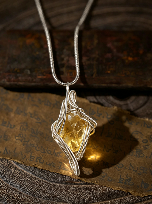 A silver wire-wrapped pendant necklace featuring a faceted yellow gemstone, displayed on a textured brown surface with a piece of aged, script-covered paper.