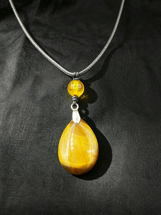 A teardrop-shaped yellow tiger's eye pendant necklace with a small round yellow bead and silver-toned metal accents, displayed on a black textured fabric background.