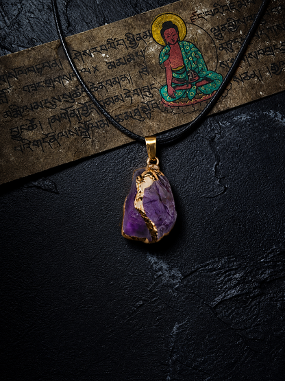 A raw purple amethyst crystal pendant with gold leaf accents, suspended on a black cord necklace, displayed against a dark textured background with a Tibetan scripture scroll and Buddha illustration.