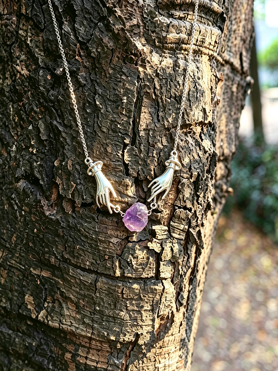 A silver-toned pendant necklace featuring two hands reaching toward a small purple amethyst stone, displayed against the textured bark of a tree outdoors.