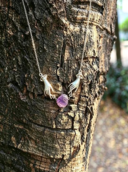 A silver-toned pendant necklace featuring two hands reaching toward a small purple amethyst stone, displayed against the textured bark of a tree outdoors.