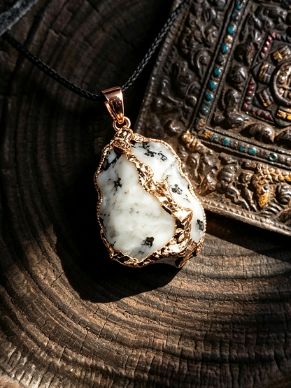 A raw white stone pendant with black inclusions, set in a gold-toned bezel with a black cord, displayed on a dark wood slice next to an ornate bronze tray.