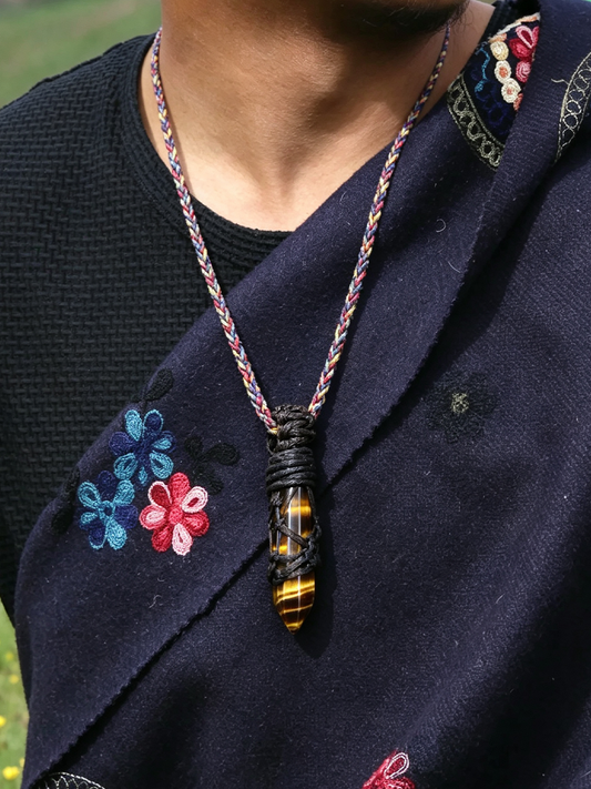 A close-up of a person wearing a braided cord necklace with a carved pendant. The pendant features a black and yellow tiger's eye pattern, and the necklace is worn over a dark navy embroidered garment.
