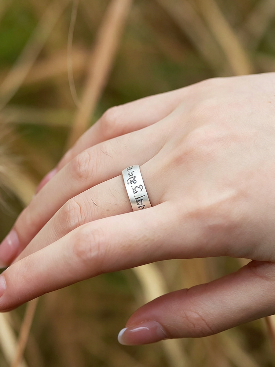 A close-up of a hand wearing a silver band ring with engraved script, set against a blurred natural background of dry grass.