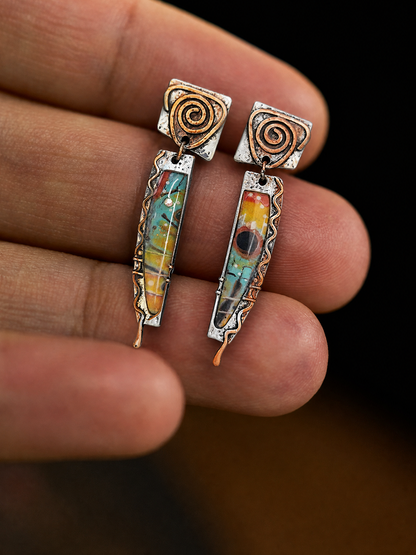 A pair of handmade dangle earrings held between fingers. The earrings feature textured silver and copper spiral tops with colorful glass inlay drops in shades of blue, yellow, orange, and green.
