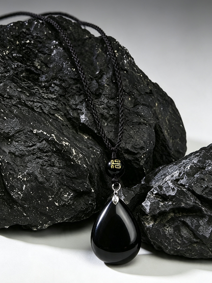 A black teardrop pendant necklace with a black braided cord, resting on dark textured rocks against a light gray background.