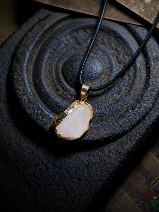A raw, irregular-shaped white crystal pendant with gold-rimmed edges, suspended on a black cord necklace, displayed on a dark textured stone surface.